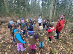 Bild 1 – Klasse 2a im winterlichen Wald Schülerinnen und Schüler der Naturpark-Grundschule Dobel stehen mit Entdeckerwesten im Wald um Waldpädagogin Regine Schirmer und hören ihren Erklärungen zu.