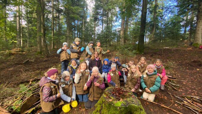 Die Naturpark-Entdecker der Grundschule Dobel stehen gemeinsam mit ihrer Klassenlehrerin im herbstlichen Wald. Die Kinder tragen Naturpark-Westen und präsentieren sich rund um einen selbstgebauten Kobel.